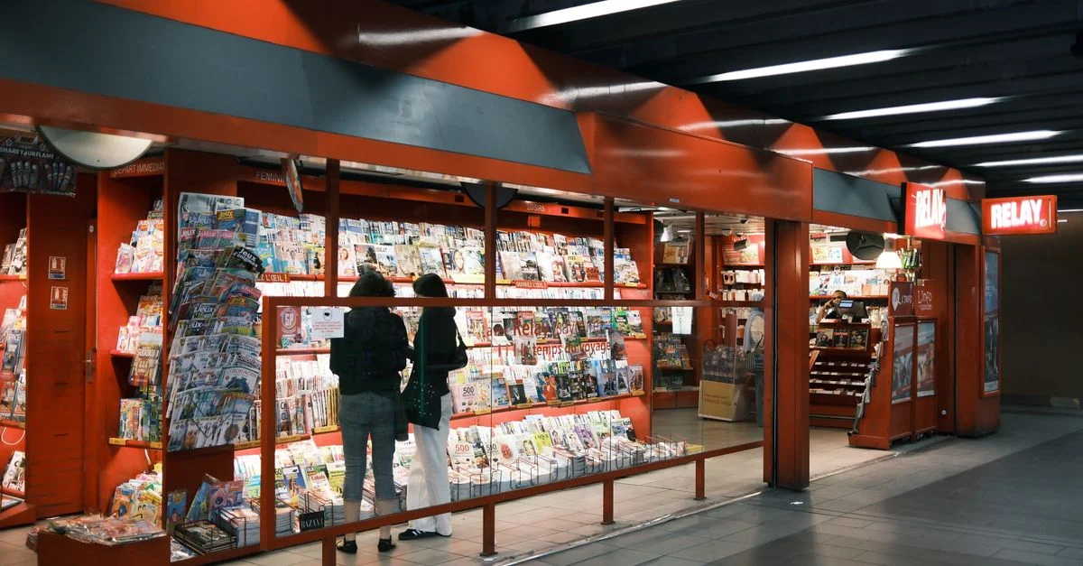 Deux adultes parcourent un kiosque à journaux rempli de magazines et de journaux dans un cadre intérieur moderne.