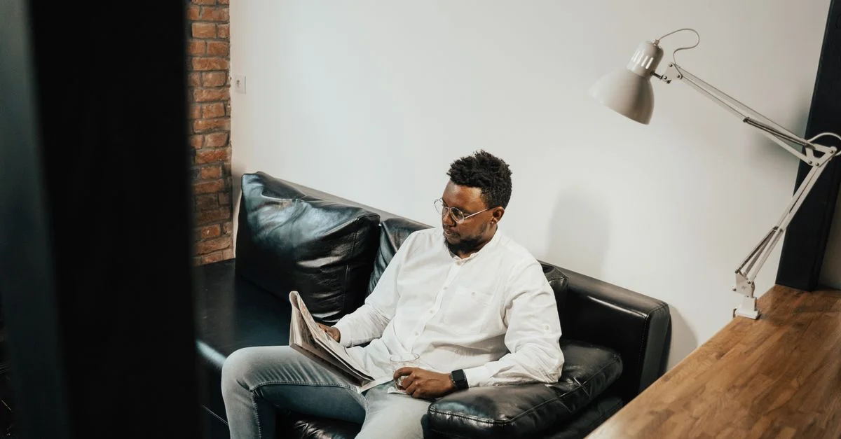 Homme se relaxant sur un canapé en cuir noir tout en lisant un journal dans un cadre intérieur moderne.