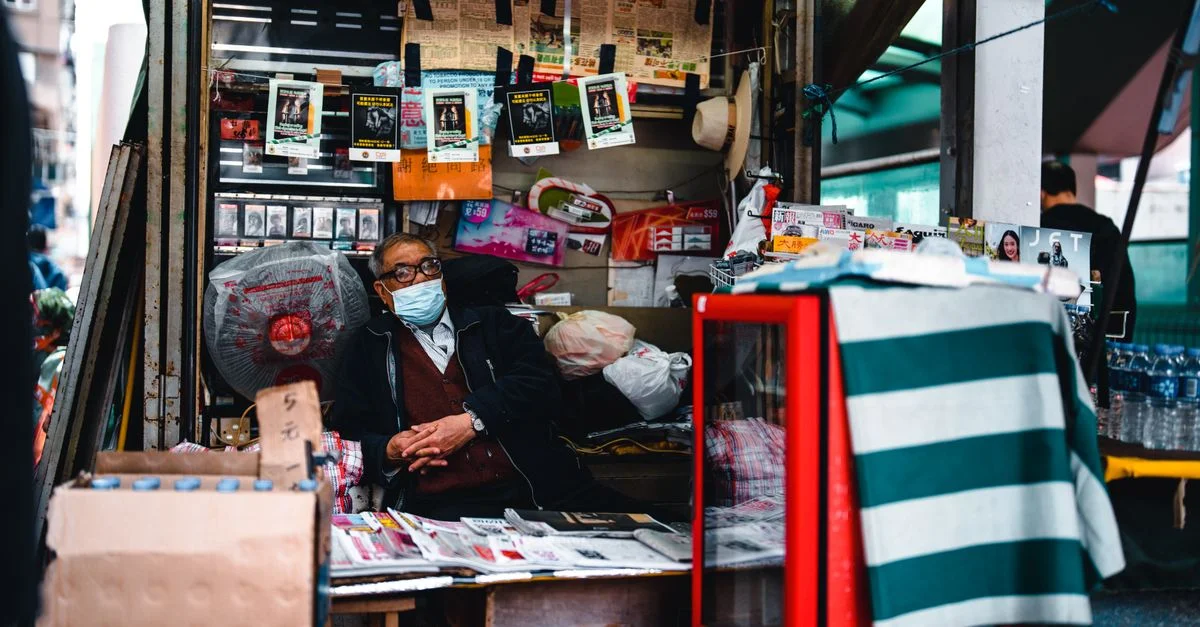 Homme âgé portant un masque gérant un kiosque à journaux animé dans un cadre urbain, promouvant la sécurité.
