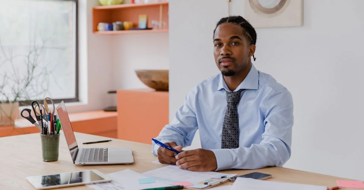Un homme d'affaires professionnel avec une barbe et des cheveux tressés est assis à son bureau avec un ordinateur portable et des documents, travaillant assidûment.
